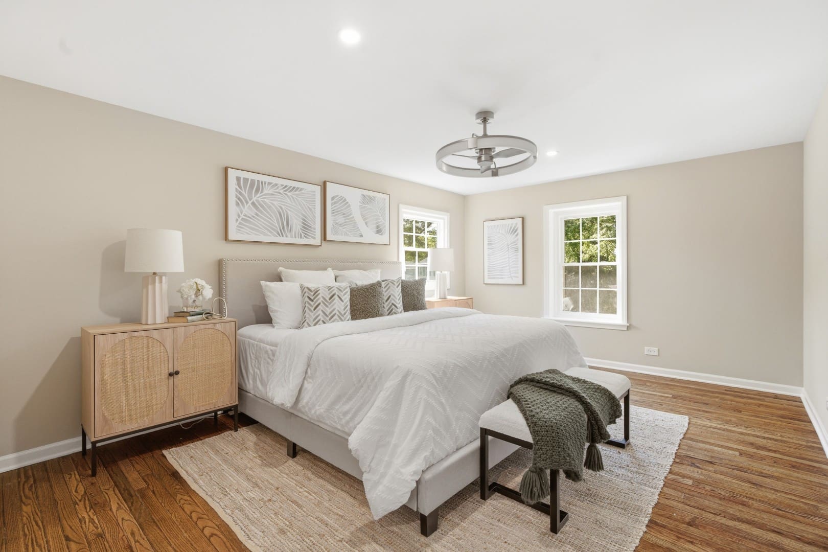 HD_1783636956552_015_primary_bed_020_1342_Winchester_Ct_Large (1) Primary bedroom featuring a beige upholstered king bed with white bedding, natural cane nightstands, neutral framed botanical art, and a modern fandelier ceiling fan.